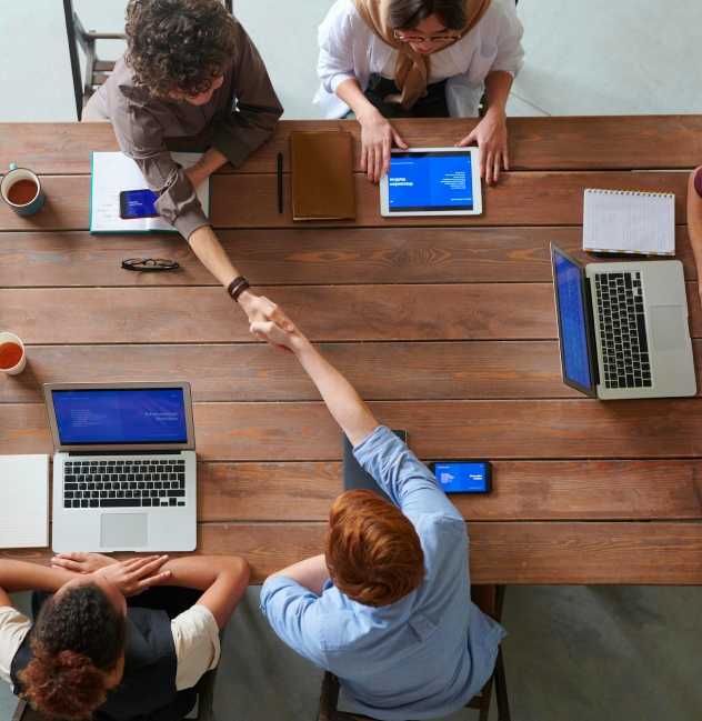 Group of business professionals sitting around a wooden table, engaged in a collaborative meeting with laptops and tablets, while two team members shake hands, symbolizing Solavita's partnership and commitment to sales cooperation